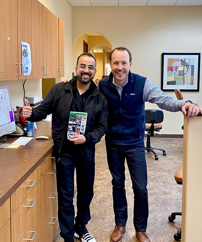 Two men smiling in orthodontic office, one holding product box, standing by desk.