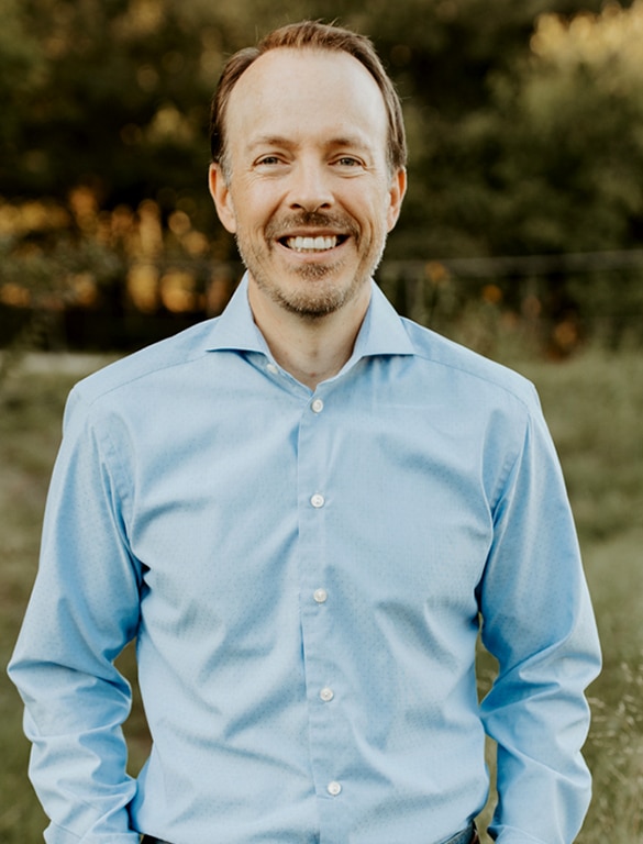 Professional portrait of man in light blue shirt smiling outdoors with green foliage background.