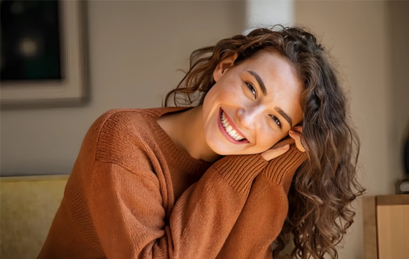 Woman smiling warmly while leaning against couch in casual home setting, wearing rust-colored sweater.