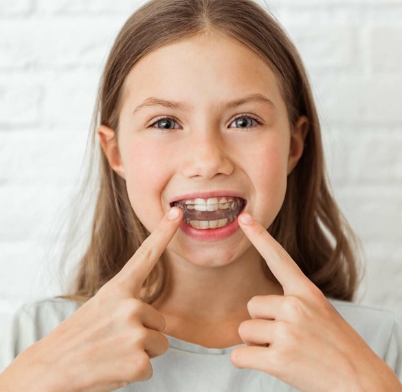 Young girl pointing to her smile in a bright clinical setting, displaying healthy teeth.