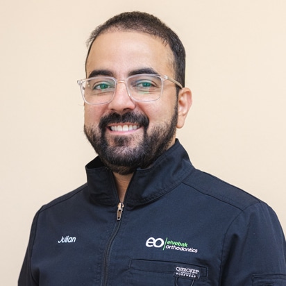 Male orthodontist wearing glasses and navy Elvebak Orthodontics jacket, smiling at camera against neutral background.